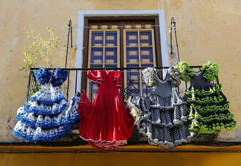 Four colorful ruffled flamenco dresses hanging on a clothesline on a balcony in front of a window.