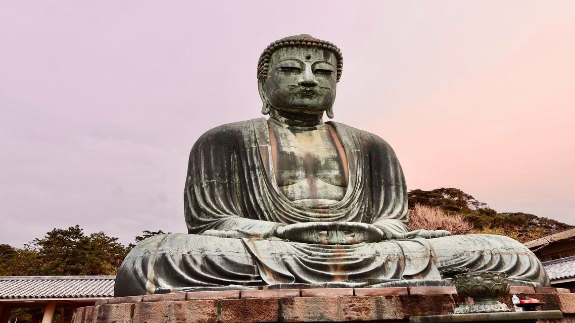 Una grande statua in bronzo patinato del Buddha si trova in posa meditativa su una base di pietra all'aperto, con alberi sullo sfondo.