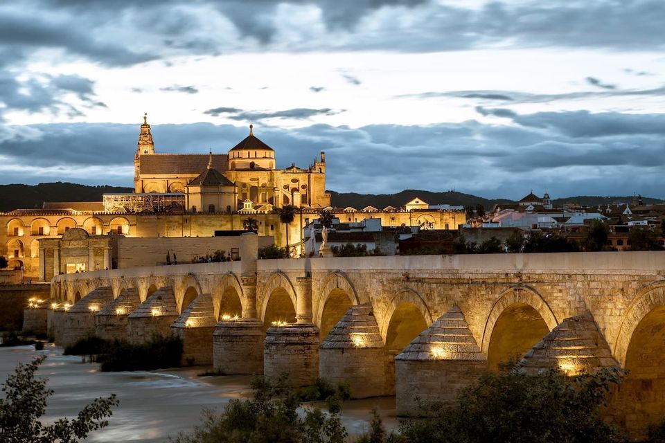 Un ponte di pietra illuminato con più arcate attraversa un fiume al tramonto, con un edificio storico sullo sfondo.