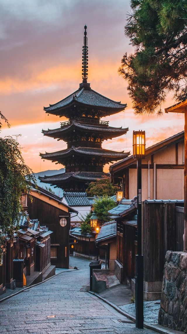Una pagoda di legno alta si erge sopra una strada di ciottoli fiancheggiata da case tradizionali e lanterne sotto un cielo al tramonto.