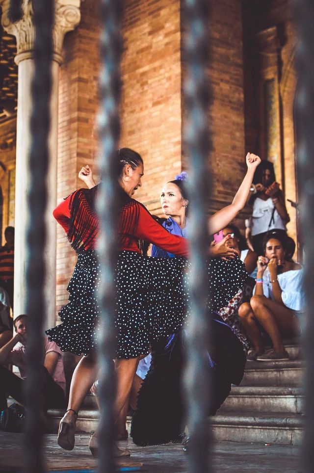 Flamenco dancers in traditional dresses perform in a stone plaza for an audience sitting on steps.