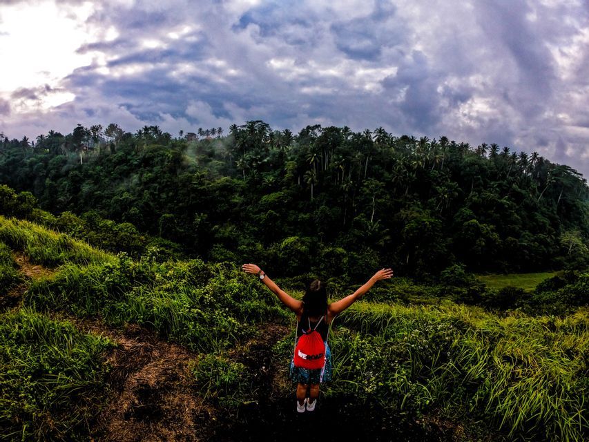 Una mujer con una mochila WeRoad roja, con los brazos extendidos en una colina de hierba, con vistas a un denso bosque verde bajo un cielo nublado.