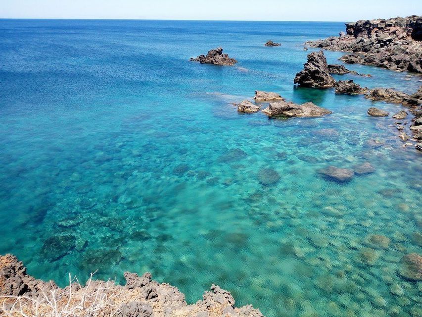 Una costa con scure rocce vulcaniche incontra il mare turchese chiaro e poco profondo, rivelando rocce sul fondale.