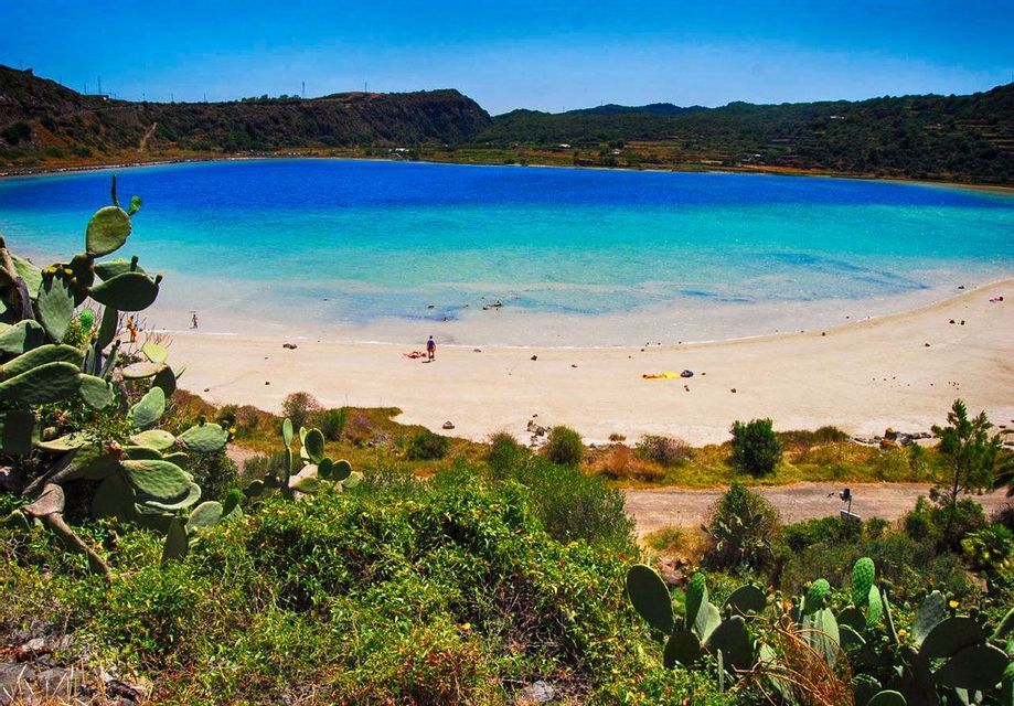Cactus di fichi d'India in primo piano si affacciano su una spiaggia di sabbia bianca e un lago azzurro bicolore circondato da verdi colline.
