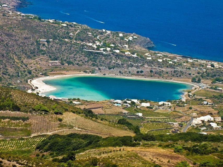 Una vista aerea di una laguna costiera circolare con acqua turchese, circondata da colline terrazzate e piccoli edifici bianchi.