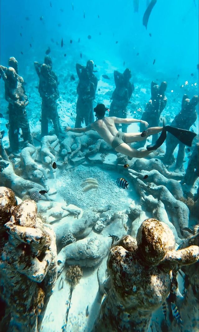 Una persona con maschera e pinne fa snorkeling sopra un'installazione circolare di statue subacquee a forma umana sul fondale sabbioso dell'oceano.