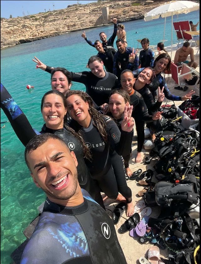 Un gruppo WeRoad sorridente in mute da sub posa per un selfie su un molo con attrezzatura da sub.