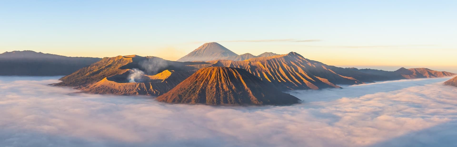 Un volcan fumant et d'autres sommets montagneux s'élèvent au-dessus d'une dense couche de nuages, illuminés par la lumière dorée du lever du soleil.
