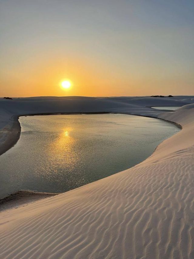 Il sole tramonta su una laguna tranquilla, proiettando la sua luce dorata su vaste dune di sabbia bianca increspata.