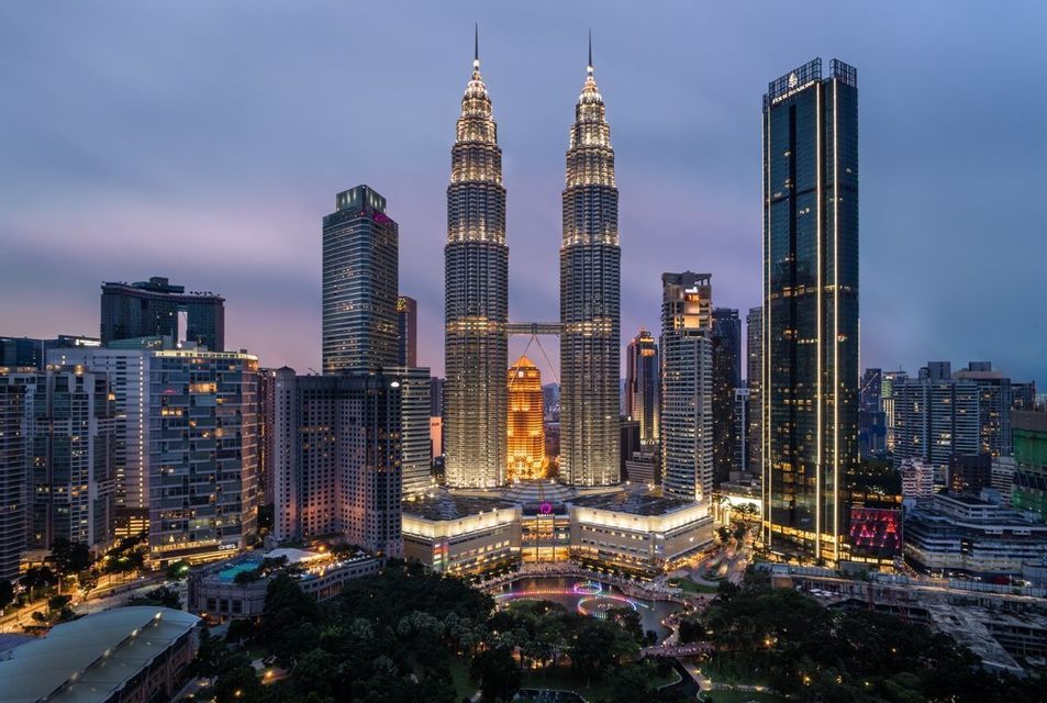 Due grattacieli gemelli illuminati con un ponte aereo dominano lo skyline di una città moderna al tramonto sotto un cielo viola.