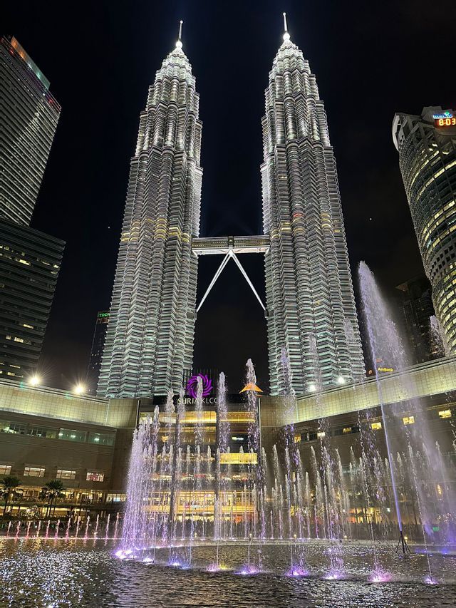Two tall, illuminated twin towers at night, with a large fountain show lit by purple and white lights in the foreground.