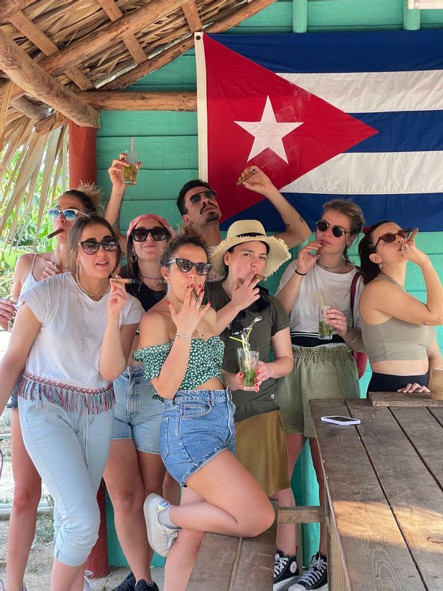 Un grupo de WeRoad posa para una foto fumando puros y bebiendo mojitos frente a una gran bandera cubana.
