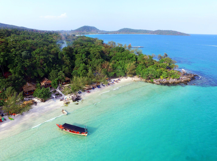 Vista aérea de una playa tropical con arena blanca, agua turquesa cristalina, un barco anclado en la costa, y bungalows entre un exuberante bosque verde.