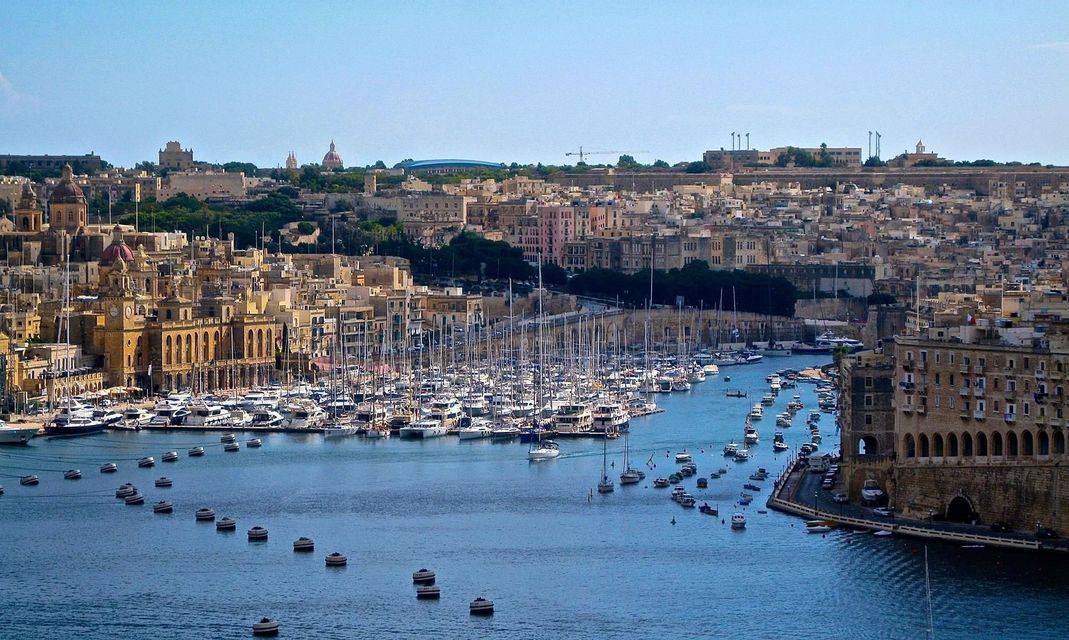 Una ciudad costera histórica con un puerto concurrido lleno de veleros atracados y otras embarcaciones bajo un cielo azul claro.