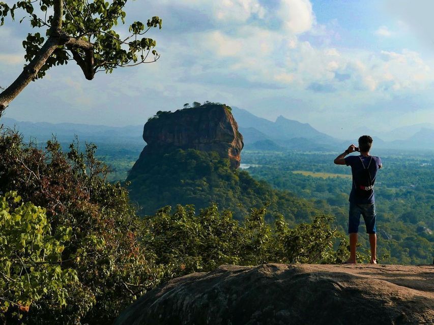 Une personne debout sur un rebord rocheux photographie une grande formation rocheuse s&#39;élevant au-dessus d&#39;un vaste paysage forestier verdoyant.