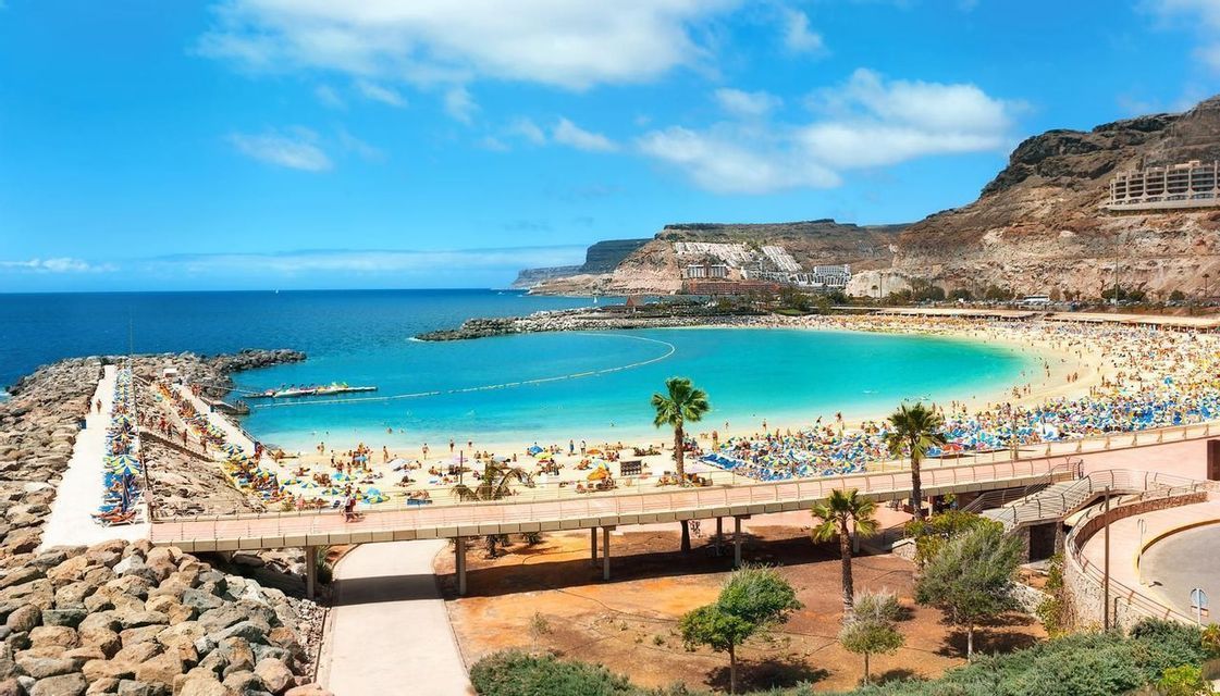 Blick von oben auf einen belebten sichelförmigen Strand mit türkisfarbenem Wasser, eingebettet in einer Bucht zwischen Felsklippen.
