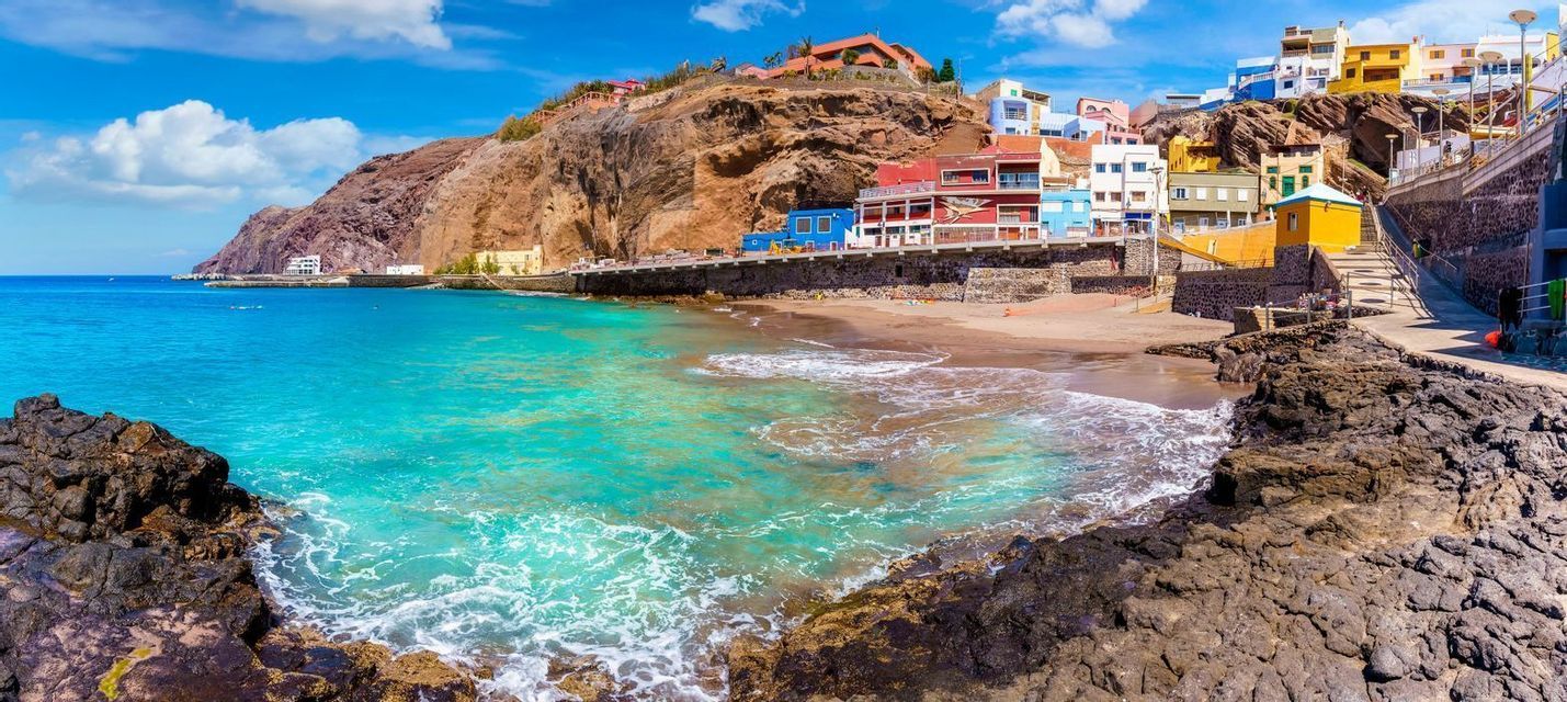 Ein Panoramablick auf ein Küstendorf mit bunten Gebäuden an einer Klippe neben einem Strand mit türkisfarbenem Wasser.