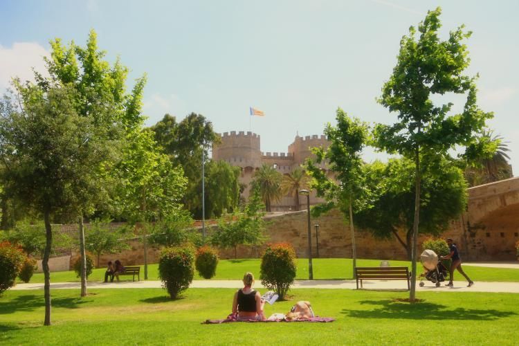 Un parco soleggiato con alberi verdi lussureggianti e persone che si rilassano, con una grande fortezza in pietra sullo sfondo.