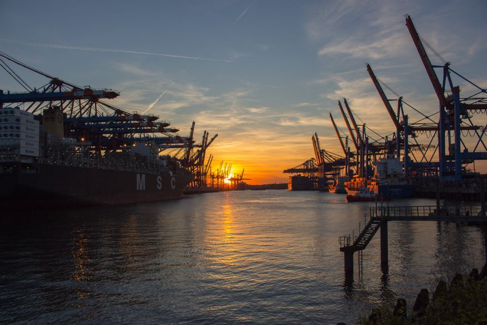 Grandi navi portacontainer e gru si stagliano in un porto industriale al tramonto, con la luce del sole che si riflette sull'acqua.