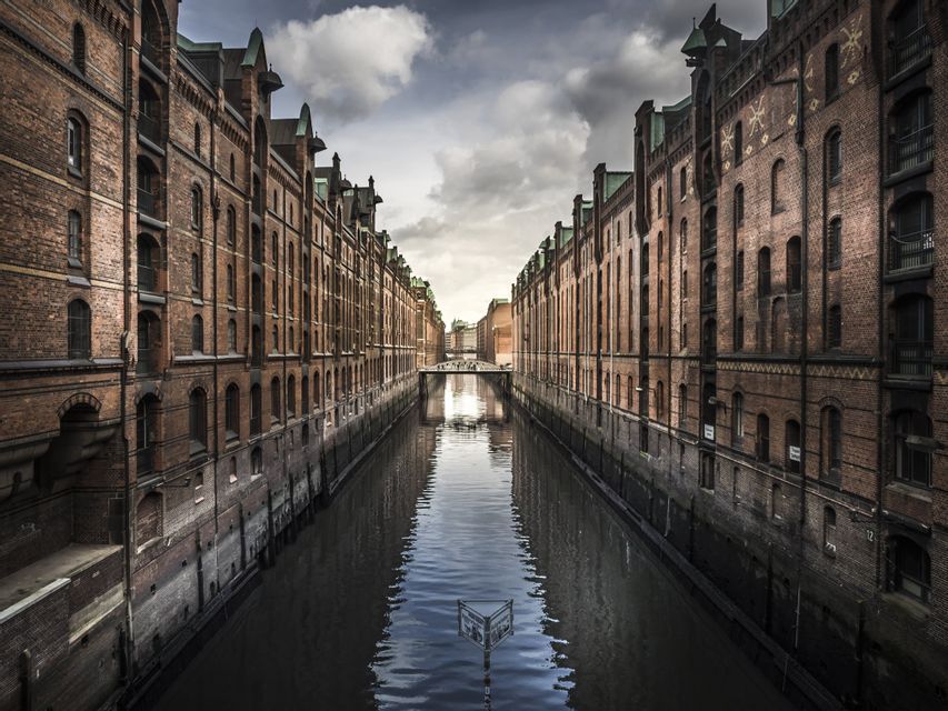 Un canale scorre tra due file di alti edifici in mattoni rossi sotto un drammatico cielo nuvoloso, con un ponte visibile in lontananza.