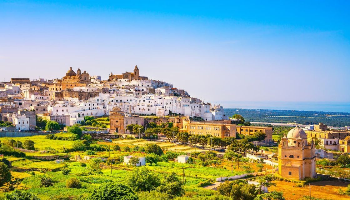 A panoramic view of a whitewashed town on a green hill overlooking the sea under a clear blue sky.