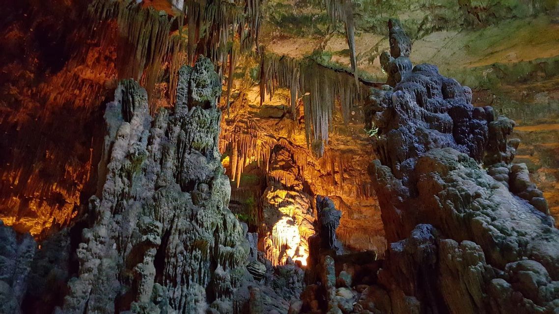 The interior of a large cave with numerous stalactites and stalagmites illuminated by warm, artificial lights.