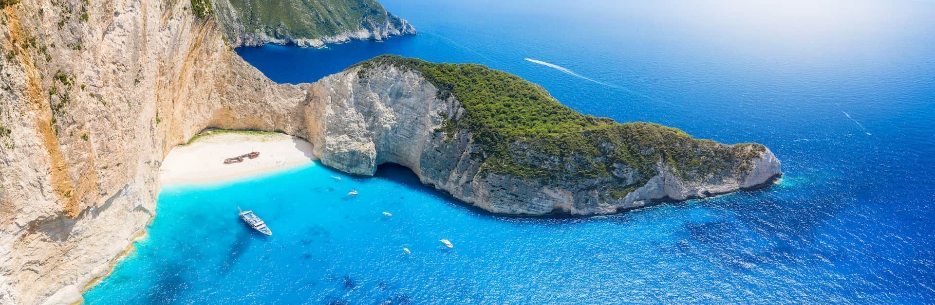 Vista aerea di un naufragio su una spiaggia isolata di sabbia bianca in una cala, circondata da scogliere e acqua turchese con barche.