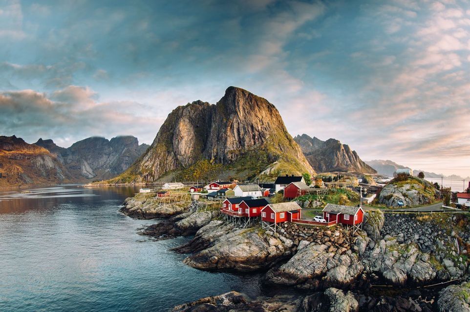A village of red and white houses on a rocky shoreline at the base of a large mountain next to a body of water.