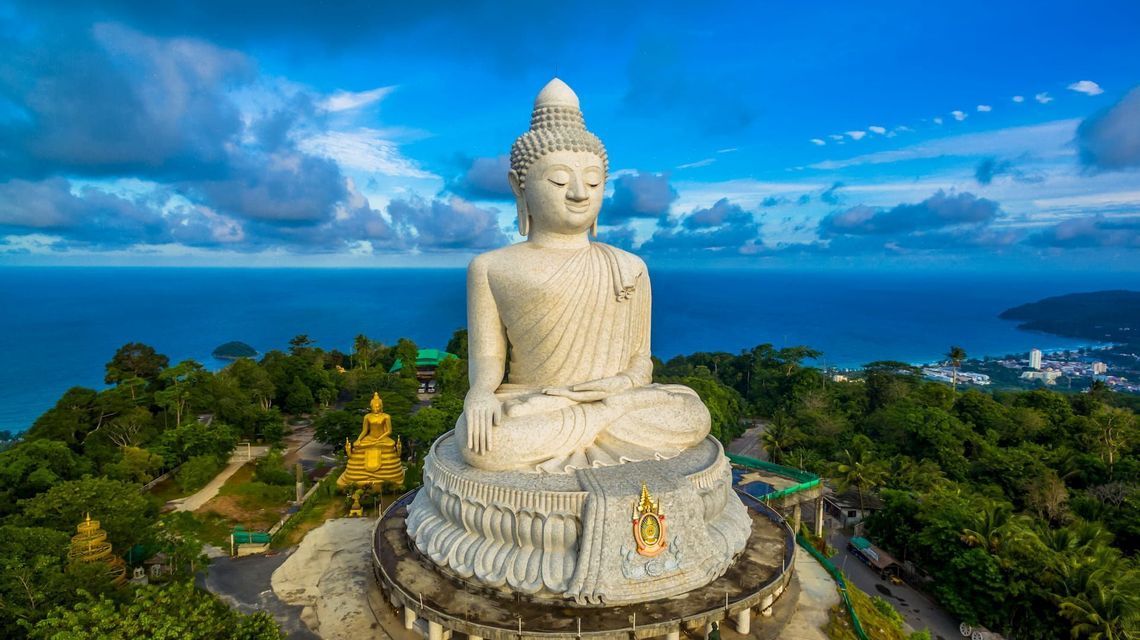 Eine große, weiße Stein-Buddha-Statue sitzt auf einem grünen Hügel mit Meerblick unter einem strahlend blauen Himmel.