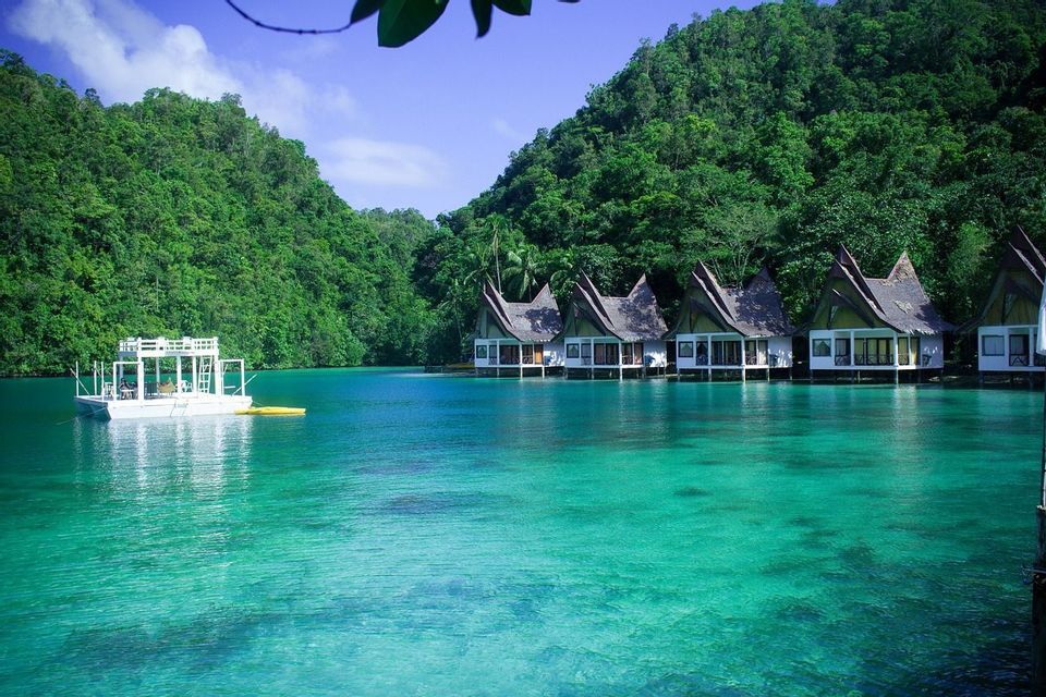 Una hilera de bungalows sobre el agua y un barco blanco en una laguna turquesa cristalina al pie de colinas exuberantes y boscosas.