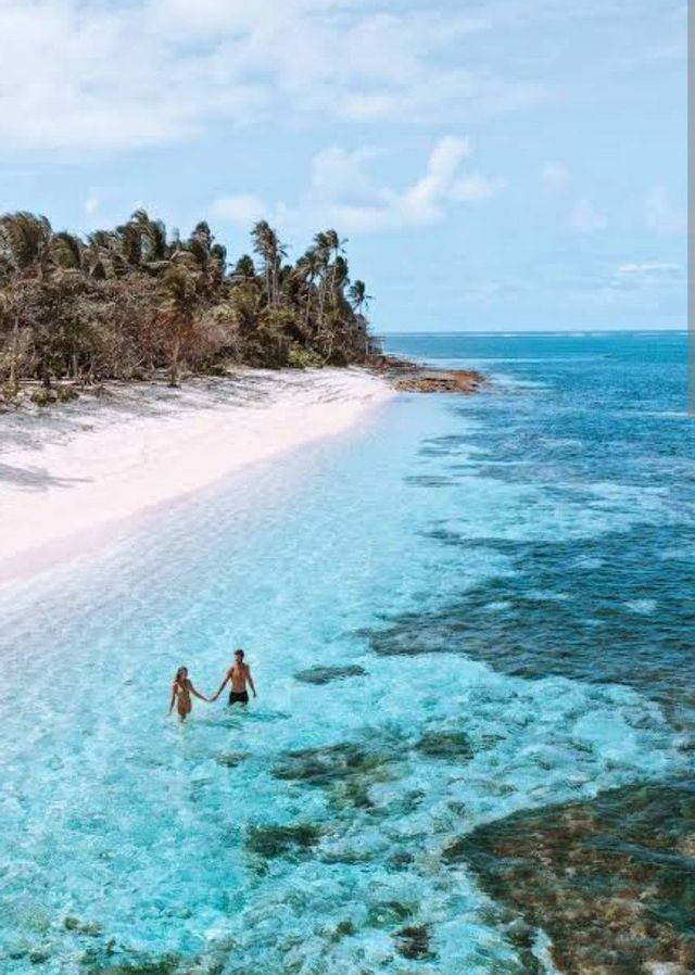 Vista aérea de una pareja cogida de la mano de pie en aguas poco profundas, claras y turquesas frente a una playa de arena blanca con palmeras.