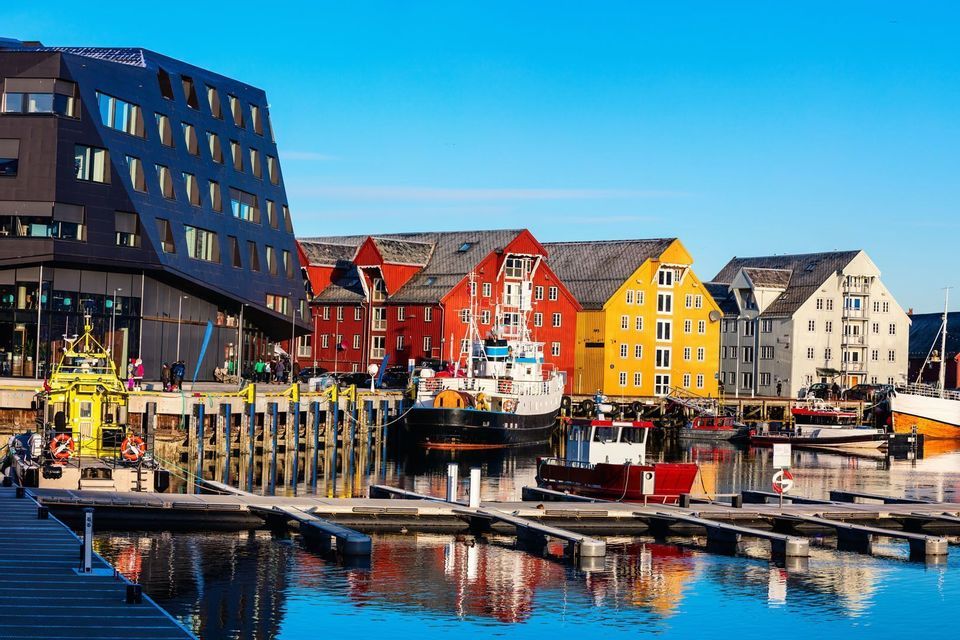 Un porto con barche ormeggiate, fiancheggiato da colorate case tradizionali e un edificio moderno, sotto un cielo blu brillante.