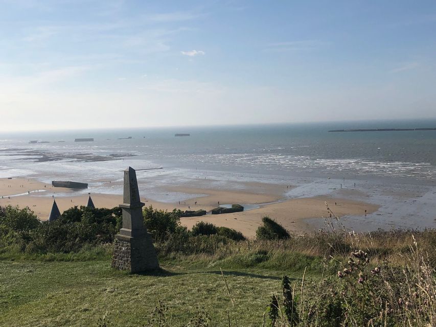 Un monument en pierre sur une colline herbeuse surplombe une plage avec des structures en béton et la mer à marée basse.