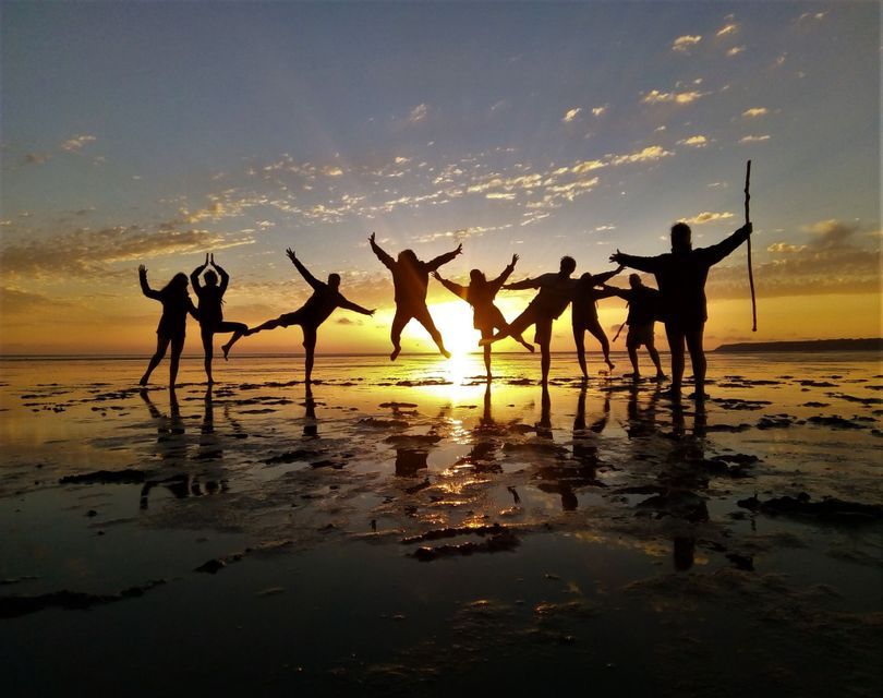 Un groupe WeRoad en voyage, silhouetté au coucher du soleil, posant joyeusement sur une plage mouillée, leurs reflets dans l'eau.