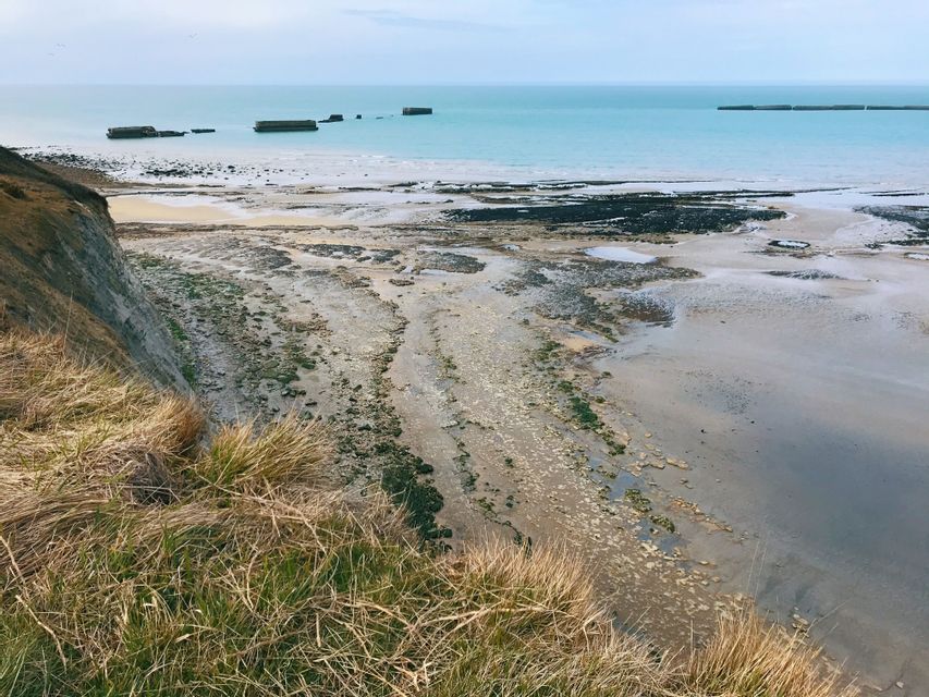 Une vue depuis une falaise herbeuse montre une plage rocheuse à marée basse et des ruines en béton dans la mer calme et turquoise.