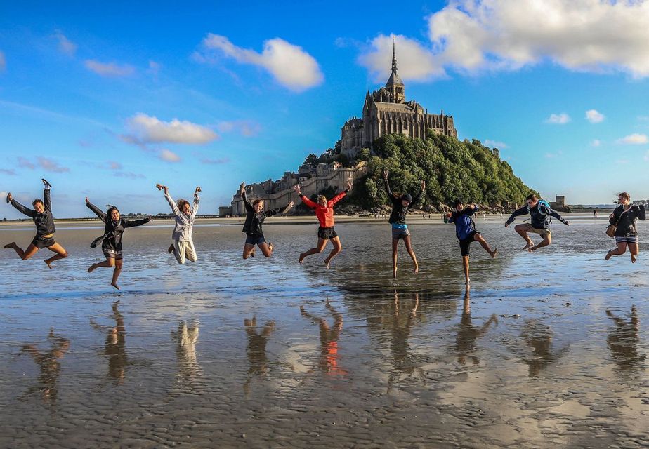 Un grupo de WeRoad salta al unísono en una playa de arena mojada, sus reflejos visibles, con un castillo en una colina al fondo.