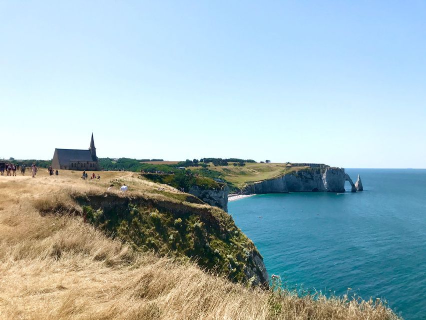 Une église se dresse sur une falaise herbeuse surplombant la mer bleue, avec une formation rocheuse en forme d&#39;arche naturelle au loin.