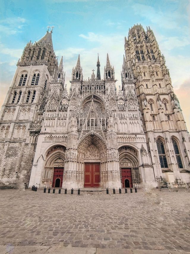 La façade ornée d&#39;une cathédrale gothique avec deux tours et des portes rouges, vue depuis une place pavée sous un ciel bleu.