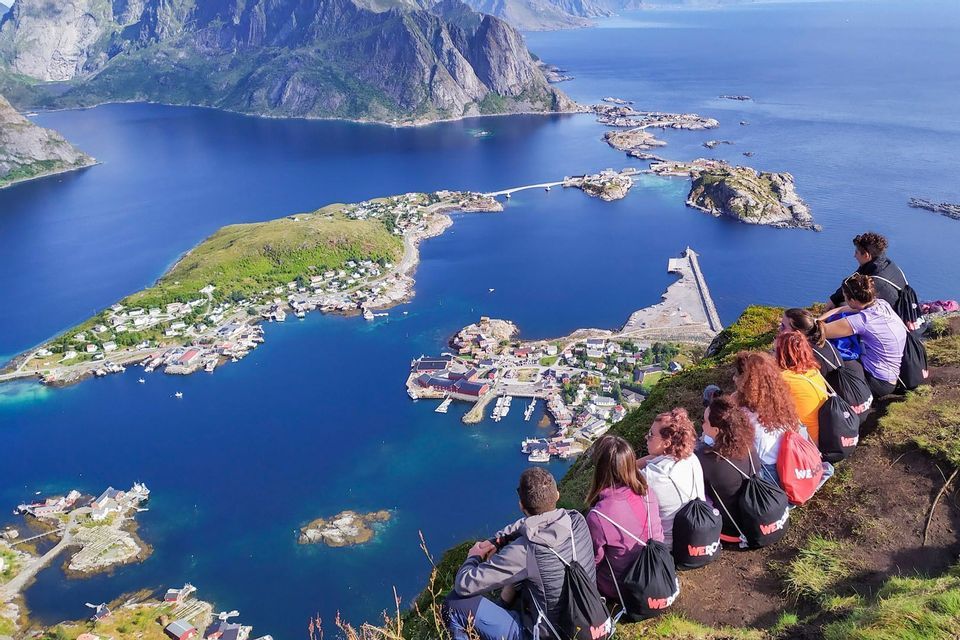 A WeRoad group trip sits on a grassy cliff, overlooking a coastal village surrounded by blue water and mountains.