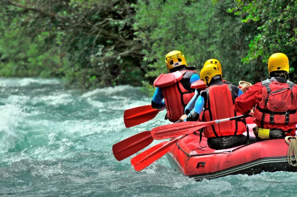 Un groupe WeRoad en voyage de rafting à bord d'un bateau gonflable rouge sur une rivière rapide, vus de dos alors qu'ils pagayent.