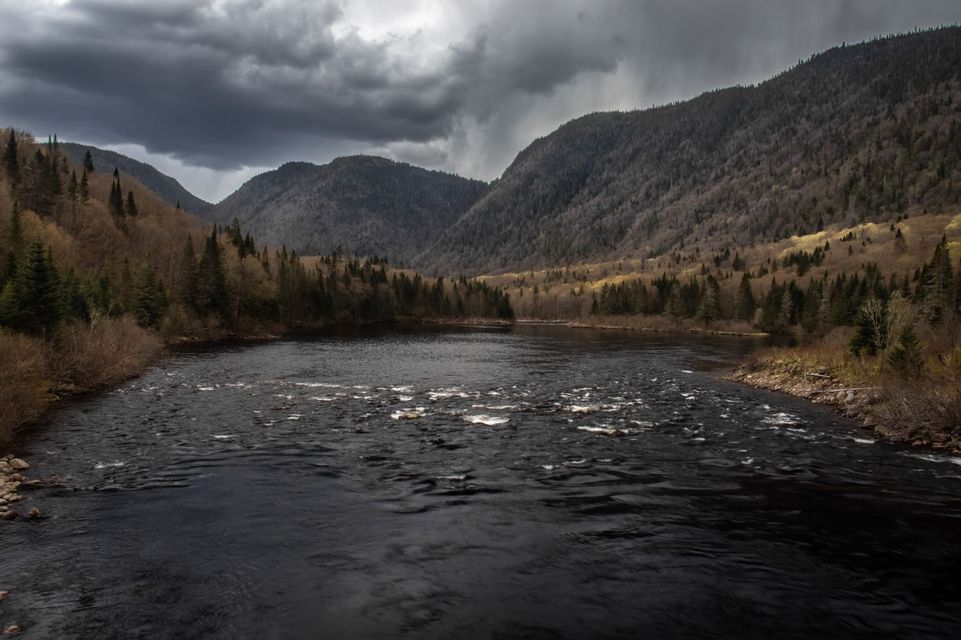 Ein dunkler Fluss fließt durch ein Tal zwischen bewaldeten Bergen unter einem Himmel voller schwerer, stürmischer Wolken.