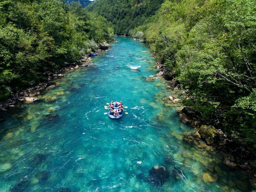 An aerial view of a WeRoad group trip rafting in an inflatable boat on a vibrant turquoise river surrounded by green forests.