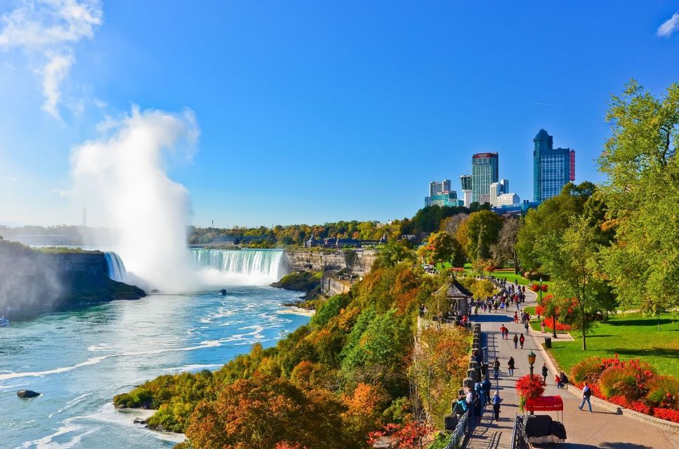 A wide waterfall generates a large cloud of mist, with people walking on a path in a park nearby and a city skyline under a blue sky.