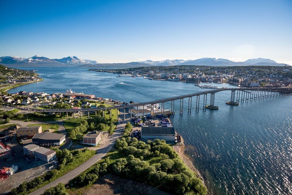 Una vista aerea di un lungo ponte che attraversa un ampio canale d'acqua verso una città costiera, con montagne innevate in lontananza.
