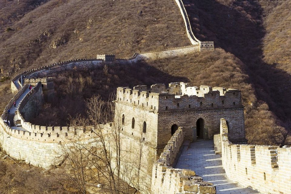 Un lungo muro di pietra con torri di guardia si snoda attraverso colline aride e marroni sotto la luce intensa del sole.