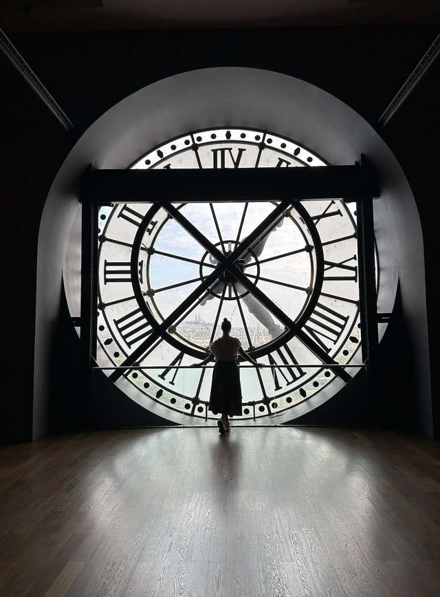 Una donna in silhouette si trova su un pavimento di legno, guardando un panorama urbano attraverso una grande finestra a orologio.