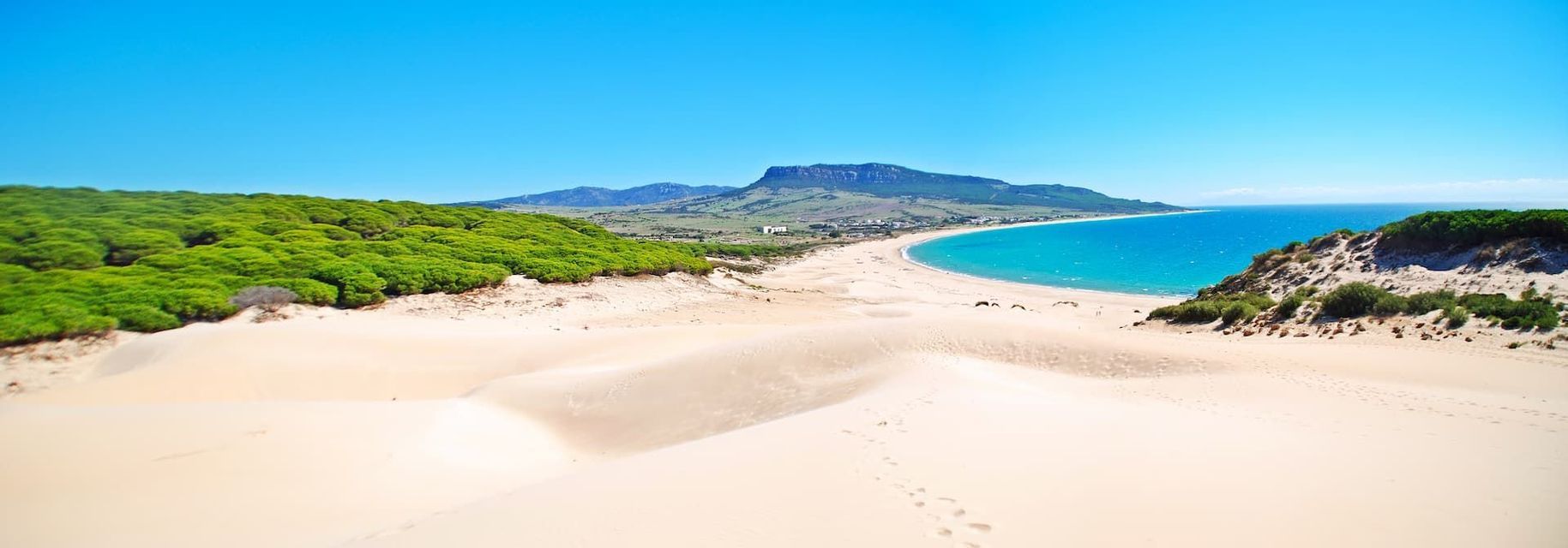 Ein Blick von oben auf weiße Sanddünen, die zu einem geschwungenen Strand mit türkisfarbenem Wasser führen, begrenzt von einem grünen Wald und fernen Bergen.