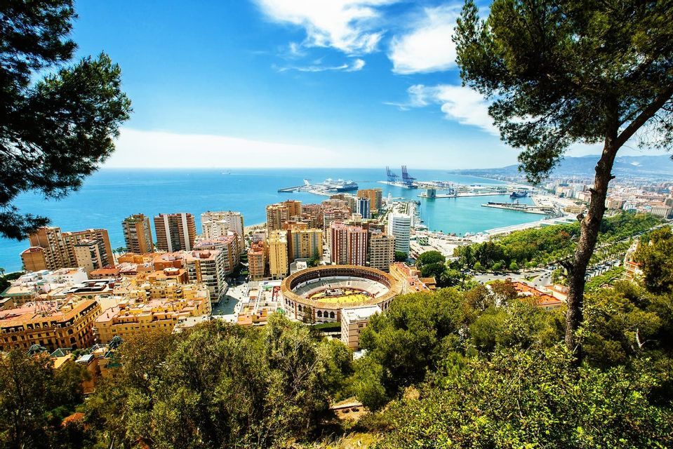 Veduta dall'alto di una città costiera con arena, un grande porto e il mare sotto un cielo azzurro.