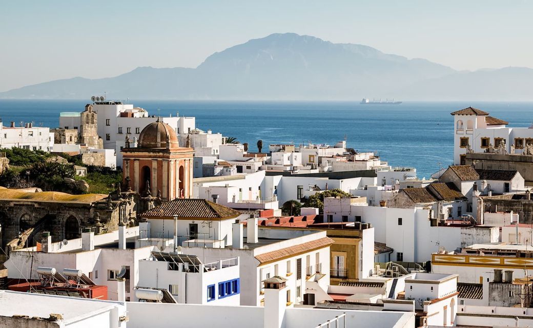Una vista dall'alto di una cittadina costiera con edifici imbiancati affacciati sul mare e una grande montagna in lontananza.