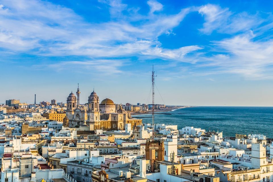 Una vista aerea di una vasta città costiera con edifici bianchi e un'imponente cattedrale a cupola vicino all'oceano sotto un cielo blu.